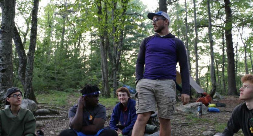 One person stands while others sit at a campsite in a wooded area. 